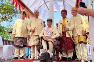 Group of men in cream traditional outfits standing on a decorated stage, with a small dog at their feet in front.