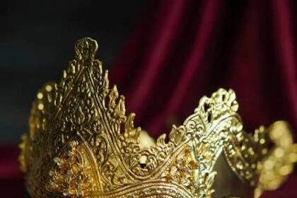 Ornate gold crown with intricate filigree resting on a dark base, surrounded by jewelry on burgundy fabric.