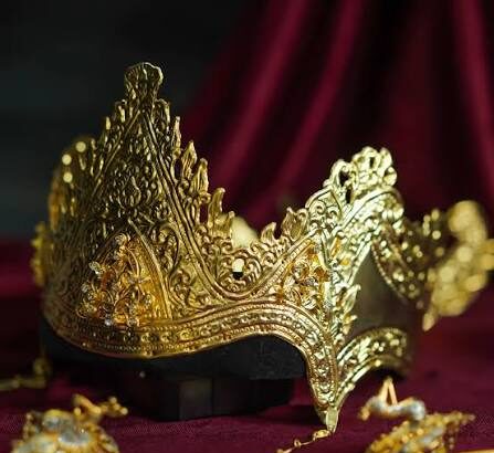 Ornate gold crown with intricate filigree resting on a dark base, surrounded by jewelry on burgundy fabric.