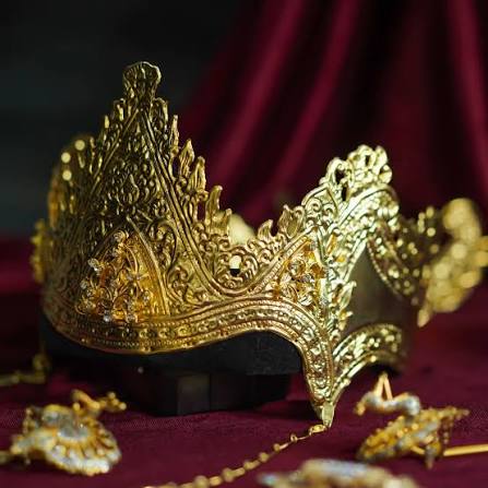 Ornate gold crown with intricate filigree resting on a dark base, surrounded by jewelry on burgundy fabric.