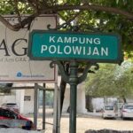 Green street sign reading 'KAMPUNG POLOWIJIAN' on a decorative post, with a banner and parked cars behind.