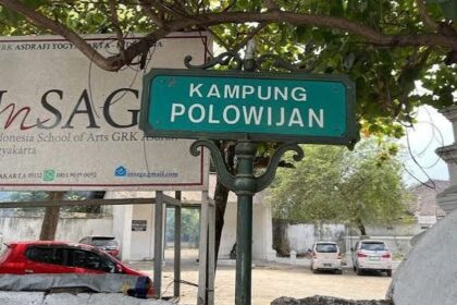 Green street sign reading 'KAMPUNG POLOWIJIAN' on a decorative post, with a banner and parked cars behind.