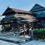 Traditional multi-tiered wooden house on stilts in a sunny garden, with a raised walkway and a lighthouse visible in the background.
