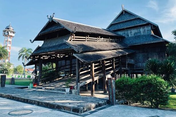 Traditional multi-tiered wooden house on stilts in a sunny garden, with a raised walkway and a lighthouse visible in the background.