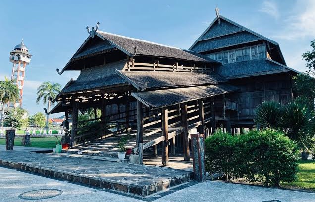 Traditional multi-tiered wooden house on stilts in a sunny garden, with a raised walkway and a lighthouse visible in the background.