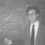 Male professor in a suit and glasses standing in front of a chalkboard full of complex equations and diagrams, pointing with chalk.