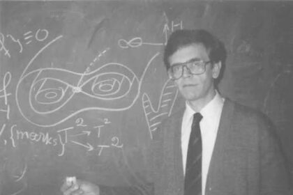 Male professor in a suit and glasses standing in front of a chalkboard full of complex equations and diagrams, pointing with chalk.