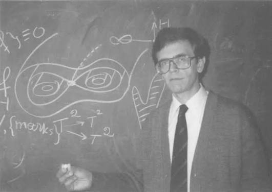 Male professor in a suit and glasses standing in front of a chalkboard full of complex equations and diagrams, pointing with chalk.