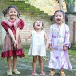 Three young girls stand on a stone patio, smiling and laughing; left girl in a red dress, middle girl in a white dress, right girl in a lavender patterned dress with pigtails.