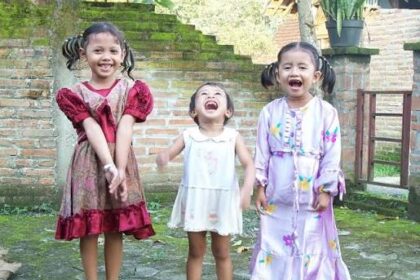 Three young girls stand on a stone patio, smiling and laughing; left girl in a red dress, middle girl in a white dress, right girl in a lavender patterned dress with pigtails.