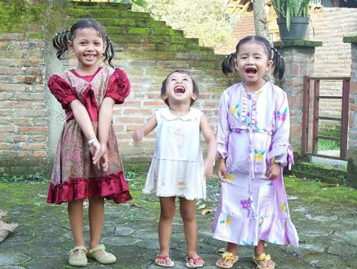 Three young girls stand on a stone patio, smiling and laughing; left girl in a red dress, middle girl in a white dress, right girl in a lavender patterned dress with pigtails.