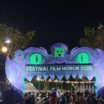 Nighttime festival entrance shaped like a purple monster with glowing green eyes; banner reads Festival Film Horor 2025, with a crowd underneath.