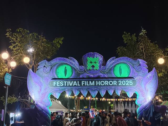Nighttime festival entrance shaped like a purple monster with glowing green eyes; banner reads Festival Film Horor 2025, with a crowd underneath.
