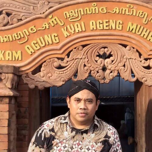 Man wearing a patterned batik shirt stands in a carved wooden entrance with gold lettering above, at a traditional Indonesian building entrance.