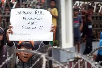 Child behind barbed wire holds a handwritten protest sign calling for immediate asset seizure, with bystanders in the background.
