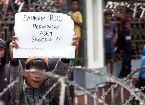 Child behind barbed wire holds a handwritten protest sign calling for immediate asset seizure, with bystanders in the background.