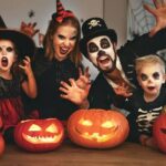 A family in Halloween costumes poses behind lit jack-o'-lanterns, with two children and two adults making spooky faces and gestures toward the camera.