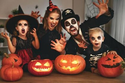 A family in Halloween costumes poses behind lit jack-o'-lanterns, with two children and two adults making spooky faces and gestures toward the camera.