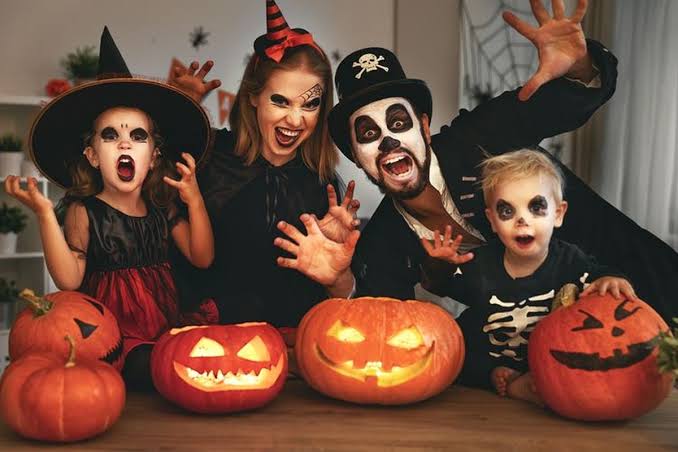 A family in Halloween costumes poses behind lit jack-o'-lanterns, with two children and two adults making spooky faces and gestures toward the camera.