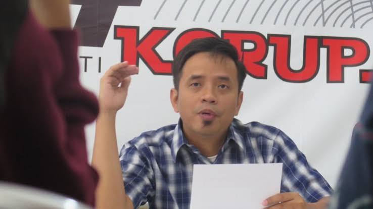 Man in a blue plaid shirt speaks into the camera while holding a sheet of paper, seated at a desk with a banner behind him that reads 'KOPRUP' (press conference scene).