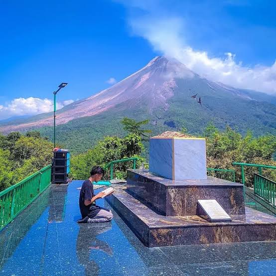A person kneels to read or pray at a marble platform and square monument on a terrace, with a volcanic mountain and blue sky in the background, and greenery around.