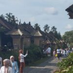 Village street with traditional thatched-roof cottages and palm trees; groups of people, many in white headscarves, walk along a cobblestone path.