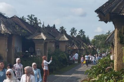 Village street with traditional thatched-roof cottages and palm trees; groups of people, many in white headscarves, walk along a cobblestone path.