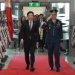 Official arrival on a red carpet: a suited dignitary walking beside a military officer who salutes, flanked by lined rifles on both sides.