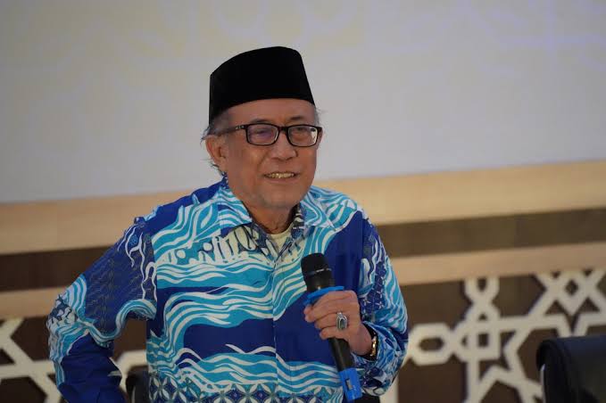 Elderly man wearing a black songkok and blue patterned shirt holds a microphone while speaking at an event