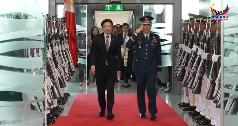 Official arrival on a red carpet: a suited dignitary walking beside a military officer who salutes, flanked by lined rifles on both sides.