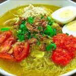 Ramen bowl with yellow broth, noodles, egg halves, tomato slices, green onions, cilantro, sesame, and red chili paste on top.