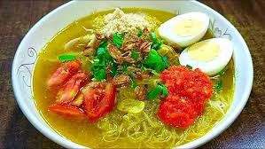 Ramen bowl with yellow broth, noodles, egg halves, tomato slices, green onions, cilantro, sesame, and red chili paste on top.