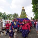 Group of people in red and purple traditional clothing marching in a festival, carrying a tall, ornamented ceremonial tree centerpiece.