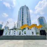 Exterior view of a large Mandiri sign with a yellow curved logo in front of tall modern buildings.