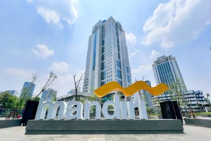 Exterior view of a large Mandiri sign with a yellow curved logo in front of tall modern buildings.