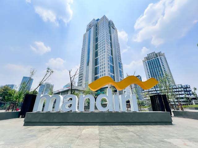 Exterior view of a large Mandiri sign with a yellow curved logo in front of tall modern buildings.