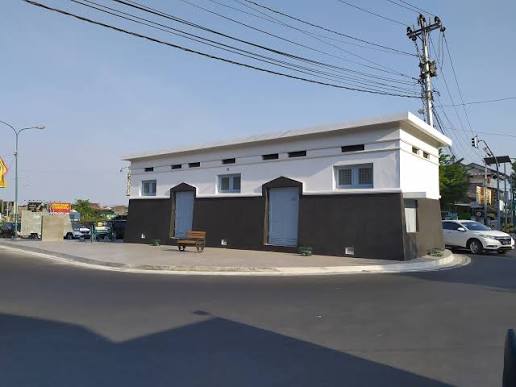 Small public utility building at a roundabout with two entrance doors and benches outside, white upper façade and dark lower wall under a clear sky.