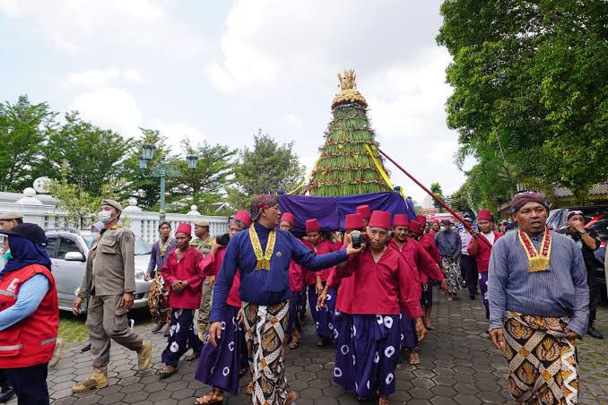 Group of people in red and purple traditional clothing marching in a festival, carrying a tall, ornamented ceremonial tree centerpiece.