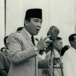 Black-and-white photo of a man in a suit and peci delivering a speech at a microphone, with helpers in the background.