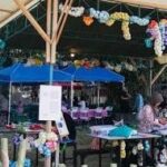 Outdoor market stall under a canopy with blue tents, grape garlands, and shoppers browsing tables in sunlight.