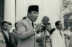 Black-and-white photo of a man in a suit and peci delivering a speech at a microphone, with helpers in the background.