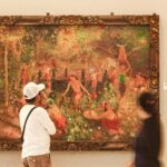 Two visitors observe a large ornate-framed painting in a museum, showing a lush garden scene with people by a water feature in the background.