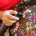 Hands engaged in stitching a dark patch onto a colorful floral batik fabric, showing careful craftsmanship.