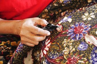 Hands engaged in stitching a dark patch onto a colorful floral batik fabric, showing careful craftsmanship.