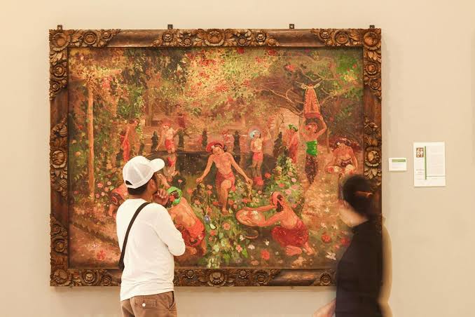 Two visitors observe a large ornate-framed painting in a museum, showing a lush garden scene with people by a water feature in the background.
