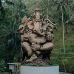 Large statue of Hindu god Ganesha with elephant head and multiple arms, seated on a pedestal in a tropical garden.