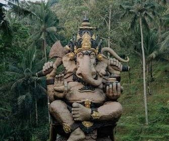 Large statue of Hindu god Ganesha with elephant head and multiple arms, seated on a pedestal in a tropical garden.