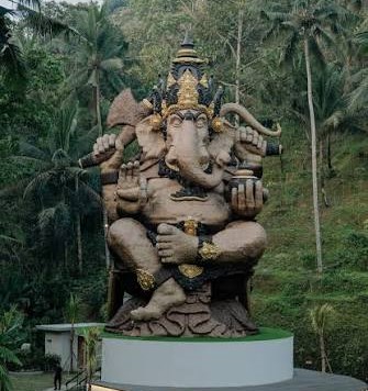 Large statue of Hindu god Ganesha with elephant head and multiple arms, seated on a pedestal in a tropical garden.