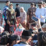 Group of people in batik attire posing at an outdoor ceremony, with audience members taking photos nearby.