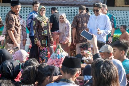 Group of people in batik attire posing at an outdoor ceremony, with audience members taking photos nearby.